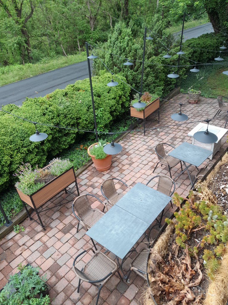 Brick-paved outdoor seating area with tables, boxes of herbs, and lights